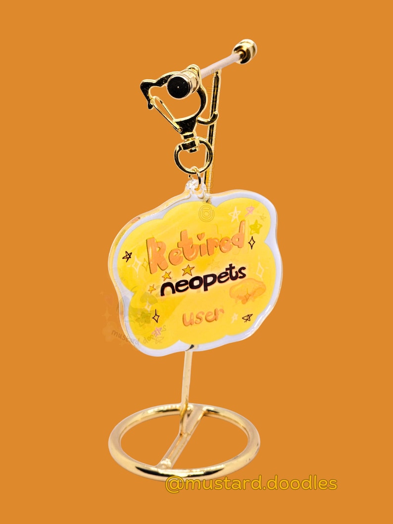 May include: A gold-colored keychain with a yellow cloud-shaped charm. The charm has the words "Retired neopets user" in orange script, with star and jelly-like graphics. The keychain is on a gold stand against an orange background.