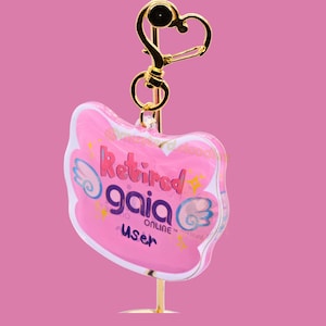 May include: A pink keychain with the text "Retired gaia online user" in purple and white. The keychain has a gold-colored heart-shaped clasp and a gold-colored stand. The background is a solid pink.