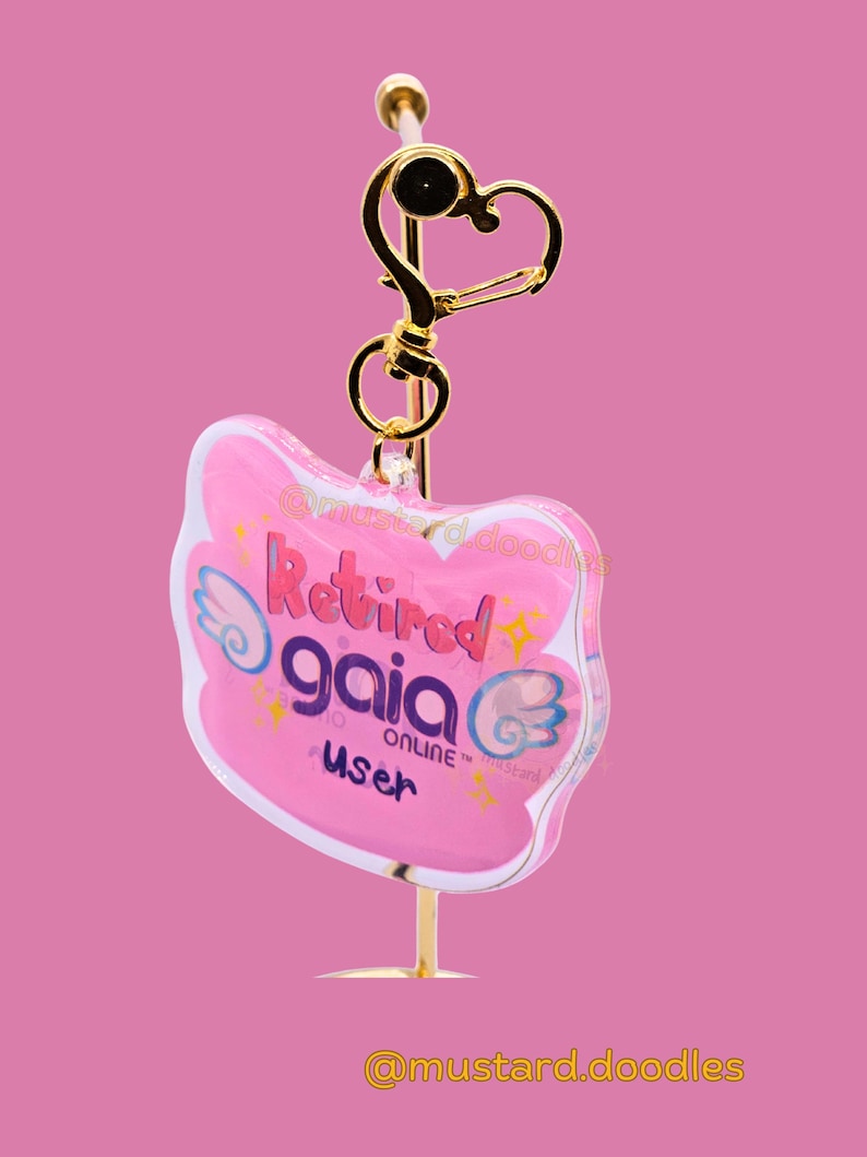 May include: A pink keychain with the text "Retired gaia online user" in purple and white. The keychain has a gold-colored heart-shaped clasp and a gold-colored stand. The background is a solid pink.