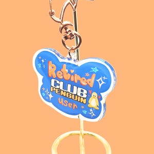 May include: A gold-colored keychain with a blue cloud-shaped charm. The charm has the words "Retired CLUB PENGUIN User" in orange and white, with a small penguin illustration. The keychain is on a gold stand against an orange background.