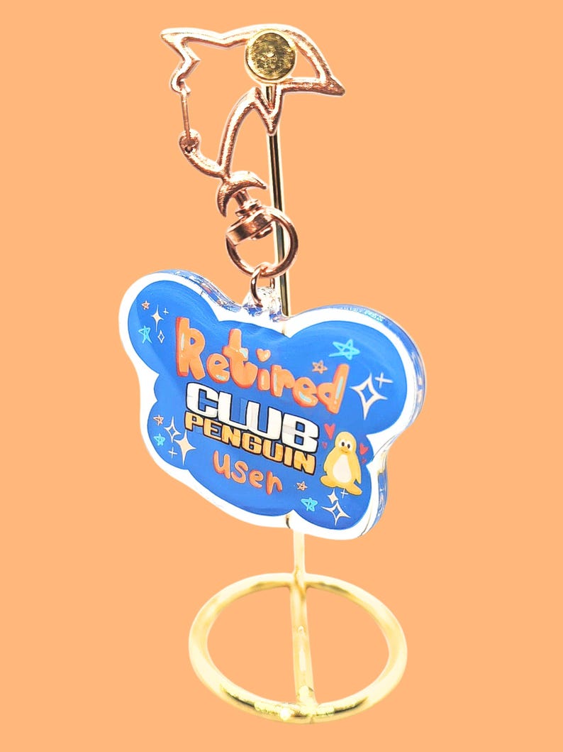 May include: A gold-colored keychain with a blue cloud-shaped charm. The charm has the words "Retired CLUB PENGUIN User" in orange and white, with a small penguin illustration. The keychain is on a gold stand against an orange background.