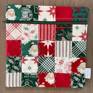 May include: A patchwork Christmas-themed zippered pouch. The design features red, green, and white squares with Santa Claus, candy canes, stars, and striped patterns. The pouch has a green zipper and a small tag with text.
