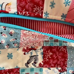 May include: Close-up of a patchwork fabric with a winter theme. The design features squares of red, white, and blue fabric with snowflakes, snowmen, and children playing in the snow. A light blue zipper is visible.