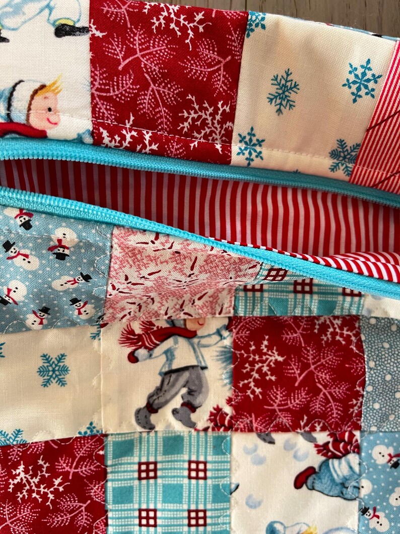 May include: Close-up of a patchwork fabric with a winter theme. The design features squares of red, white, and blue fabric with snowflakes, snowmen, and children playing in the snow. A light blue zipper is visible.