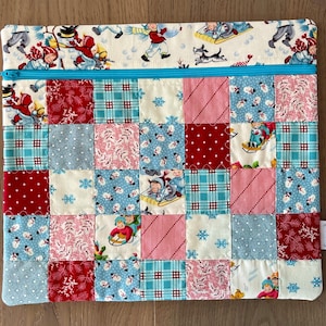 May include: A patchwork zippered pouch featuring a variety of fabrics in red, blue, and white. The top section has a cream-colored fabric with a winter scene print. The main body is a patchwork of squares with various patterns, including snowflakes and polka dots.