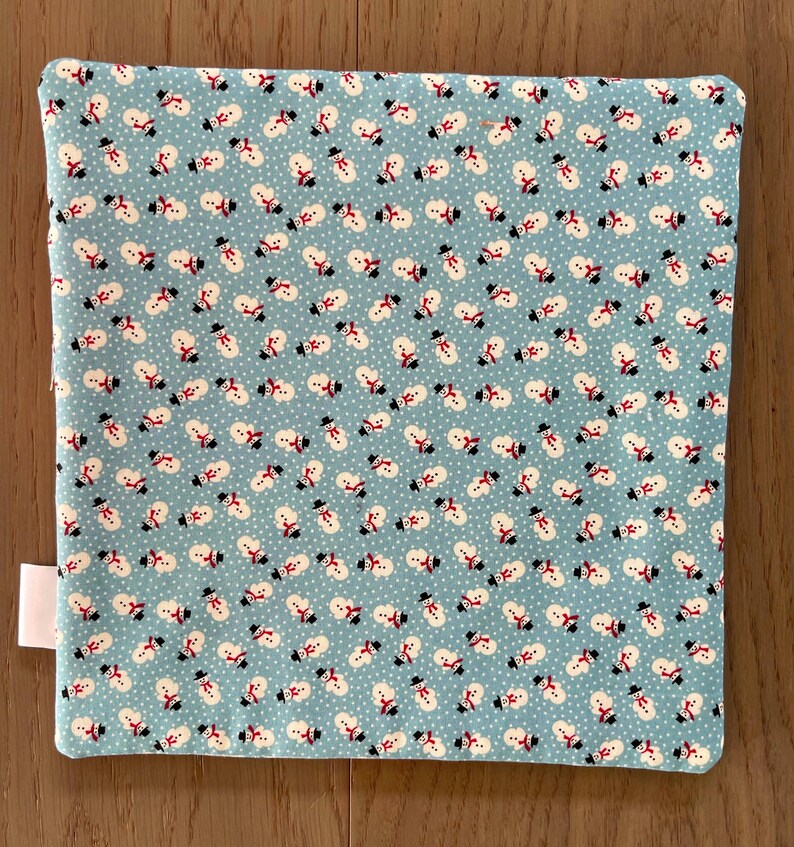 May include: A square, light blue fabric with a repeating pattern of snowmen wearing black top hats and red scarves. The snowmen are set against a background of white dots. A white tag is attached to the side of the fabric.