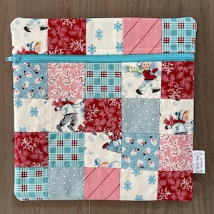 May include: A patchwork zippered pouch featuring a winter theme. The design incorporates squares of fabric with patterns of snowflakes, snowmen, and children in winter attire. The pouch has a light blue zipper and a small tag on the side.