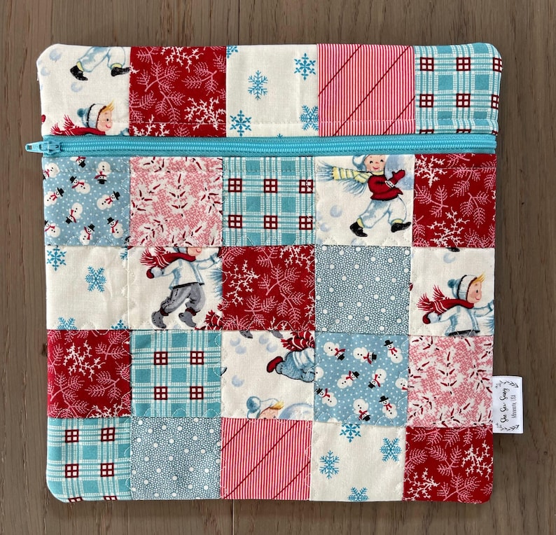May include: A patchwork zippered pouch featuring a winter theme. The design incorporates squares of fabric with patterns of snowflakes, snowmen, and children in winter attire. The pouch has a light blue zipper and a small tag on the side.