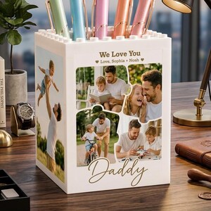 Peut inclure: Un pot à crayons blanc avec un collage de photos de famille et le mot "Daddy" en lettres dorées. Le pot contient plusieurs stylos de couleurs pastel. Le texte "We Love You" et des noms sont également visibles.