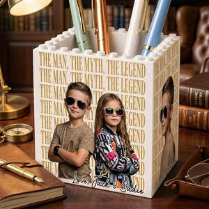 May include: A white pen holder with the text "THE MAN, THE MYTH, THE LEGEND" and photos of two children. The holder contains four pens in pastel colors. A desk lamp, notebook, and books are in the background.