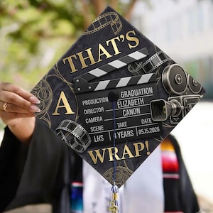 Könnte beinhalten: Ein schwarzer Doktorhut-Aufsatz mit goldenem Schriftzug THAT'S A WRAP! und einer Filmklappen-Grafik mit dem Text PRODUCTION ELIZABETH CANON, SCENE 4 YEARS, DATE 05.15.2026. Er ist mit Filmrollen-Motiven und einer blauen Quaste versehen.