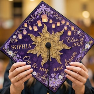 Könnte beinhalten: Ein lila Abschluss-Hut mit goldenen Akzenten, der ein Sonnendesign und den Namen "Sophia" zeigt. Der Hut ist mit floralen Elementen, Laternen und dem Text "Class of 2026" und "On to my new dream" verziert.