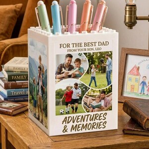 Könnte beinhalten: Ein weißer Stifthalter mit einer Collage von Familienfotos und dem Text "FOR THE BEST DAD FROM YOUR SON, LEO" und "ADVENTURES & MEMORIES." Er enthält fünf Stifte in Pastellfarben. Eine gerahmte Zeichnung und Bücher sind im Hintergrund.
