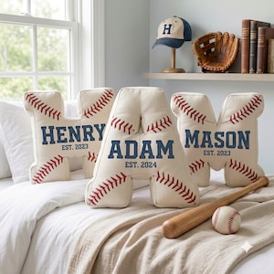 Puede incluir: Tres cojines con forma de letra hechos de tela color hueso con costuras rojas estilo béisbol. Los cojines presentan los nombres Henry, Adam y Mason en letras azules, junto con el texto EST. 2023 o EST. 2024 impreso debajo de cada nombre.