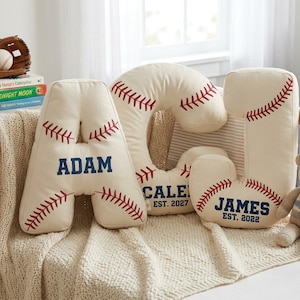 Peut inclure: Trois coussins en forme de lettres de couleur crème conçus pour ressembler à des balles de baseball avec des coutures rouges. Les coussins portent les noms ADAM, CALEB et JAMES en bleu, avec EST. 2027 et EST. 2022 imprimés sous les noms.