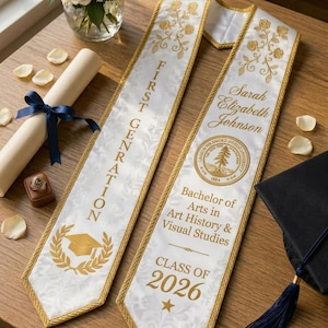 May include: A white graduation stole with gold embroidery and trim. The left side features the text FIRST GENRATION and a graduation cap icon. The right side displays the name Sarah Elizabeth Johnson, a university seal, degree text, and 2026.