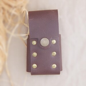 May include: A dark brown leather pouch featuring a fold-over flap secured by a single central metal snap button. The front panel is reinforced with six small, circular brass-toned metal rivets arranged in two vertical rows of three on each side.