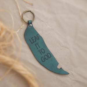 May include: A teal leather keyring shaped like a leaf with the text LEAF IT TO GOD embossed in black. The keyring features a dark metal ring attached to the top with a small metal rivet, creating a functional accessory for holding keys.