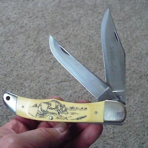 May include: A folding pocket knife with a cream-colored handle featuring an engraved image of a bear. The knife has two blades, one with the inscription "Schrade+."
