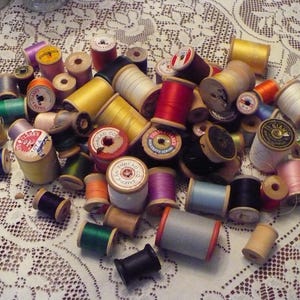 May include: A collection of wooden spools of thread in various colors, including red, blue, green, white, and brown. The spools are arranged in a pile on a white lace tablecloth.