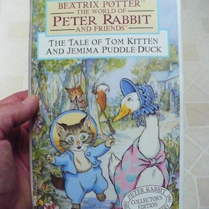 May include: A vintage VHS video cassette featuring Beatrix Potter's The Tale of Tom Kitten and Jemima Puddle-Duck. The cover art depicts a cartoon cat wearing a blue jacket and a duck wearing a pink and blue dress. The text "Peter Rabbit - And Friends" is printed in a white box on a blue background.