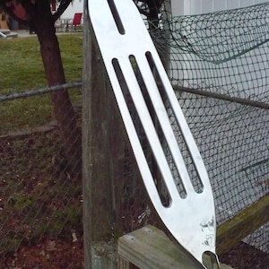 May include: A long, silver metal spatula with three long slots and one shorter slot. The spatula has a rounded end and a small hook. It is leaning against a wooden post.