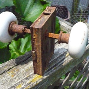May include: A rusty metal door knob with two white porcelain knobs. The knob is attached to a wooden surface.