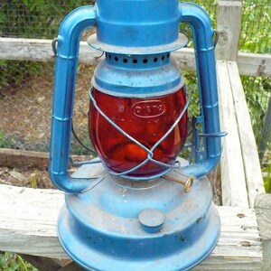 May include: Vintage blue and red lantern with a glass globe. The lantern has a blue metal frame, a red glass globe, and a handle for carrying. The lantern is sitting on a wooden surface.