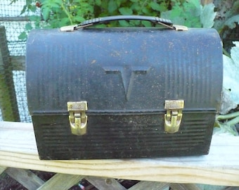 May include: Vintage black metal lunchbox with a handle and two brass-colored clasps. The lunchbox has a ribbed design and a stylized "T" emblem on the front. The lunchbox is sitting on a wooden surface.