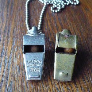 May include: Two vintage metal whistles, one silver and one brass, with a silver chain. The silver whistle is marked "THE ACME THUNDERER MADE IN ENGLAND". The whistles are on a wooden surface.