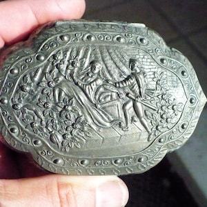 May include: A silver-toned metal trinket box with an ornate design featuring a scene of two figures in a garden setting. The box has a raised, detailed design with a floral motif.