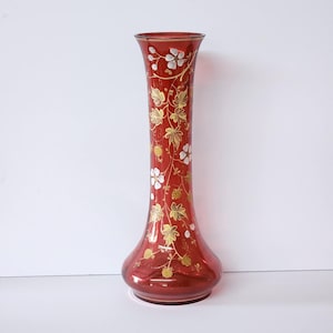 Puede incluir: Un jarrón alto de cristal rojo rubí con la parte superior acampanada y base bulbosa. El jarrón está adornado con un delicado diseño floral con hojas doradas y flores blancas. El jarrón mide unos 30 cm de altura.