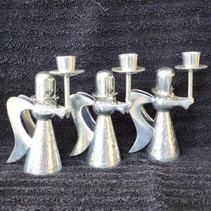 Könnte beinhalten: Set aus vier silbernen Engel-Kerzenhaltern. Jeder Engel hält einen Kerzenhalter über seinem Kopf. Die Engel stehen in einer Reihe und blicken in die gleiche Richtung. Die Kerzenhalter sind aus gehämmertem Metall und haben eine strukturierte Oberfläche.