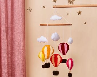 Puede incluir: Un móvil de globos aerostáticos de colores con nubes blancas que cuelgan de un anillo de madera. Los globos están hechos de fieltro y tienen rayas rojas, amarillas, naranjas y rosas.