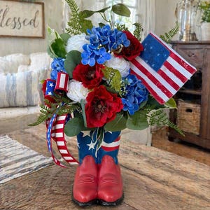 May include: A patriotic floral arrangement in a red, white, and blue color scheme. The flowers are arranged in a red cowboy boot vase with a star-spangled design. An American flag and ribbon complete the festive display.