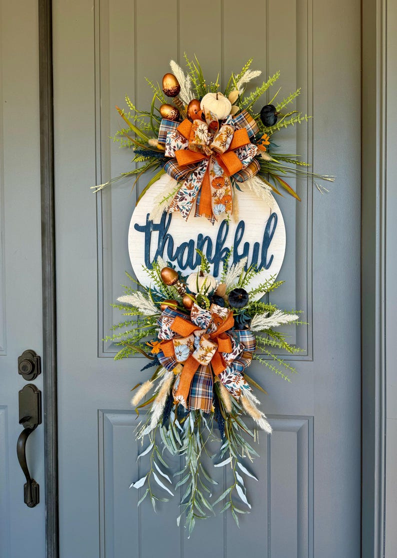 May include: A fall-themed door wreath with a round wooden sign that says 'thankful' in dark blue lettering. The wreath is decorated with orange, blue, and white ribbon, faux greenery, and small pumpkins.