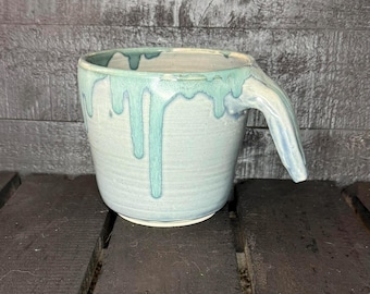 Puede incluir: Taza de cerámica hecha a mano con un diseño de asa único. La taza es de color azul claro con esmalte turquesa goteando alrededor del borde. La taza está sobre una superficie de madera oscura. Ideal para café o té.