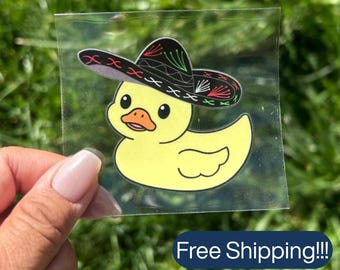 Pode incluir: Um adesivo transparente com um patinho de borracha amarelo usando um sombreiro preto com costuras decorativas vermelhas, brancas e verdes. O pato tem um bico laranja brilhante e olhos pretos, apresentado como uma ilustração gráfica num suporte quadrado.