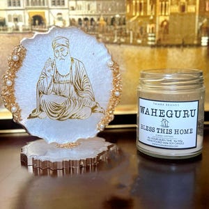 May include: A resin coaster featuring a gold-coloured religious figure, with gold trim and faux pearl embellishments. Beside it is a glass jar candle with the text "WAHEGURU BLESS THIS HOME".