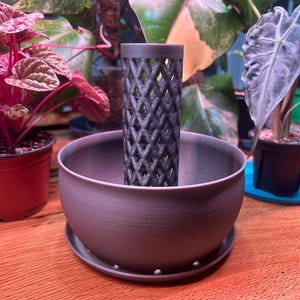 May include: A gray planter pot with a matching lattice-design insert. The pot has a wide, shallow bowl shape with a matching saucer. The insert is cylindrical and features a repeating diamond pattern. The background includes various potted plants.