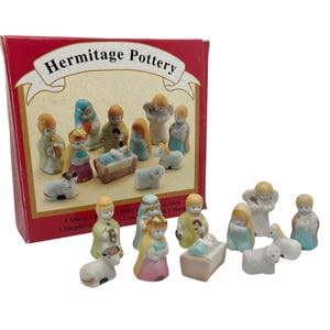 May include: A Hermitage Pottery nativity set featuring miniature figurines of Mary, Joseph, baby Jesus, wise men, angels, shepherds, sheep, and a cow. The figures are painted in soft pastel colors and come in a red box.