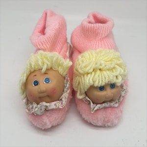 Puede incluir: Un par de calcetines de bebé de felpa rosa con caras de Cabbage Patch Kid. Las zapatillas tienen un puño de punto rosa y un ribete floral blanco alrededor de la cara.
