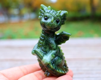 Op de afbeelding: Een klein groen jade drakenbeeldje met zwarte ogen. De draak is in een speelse stijl gebeeldhouwd en heeft vleugels en een staart.
