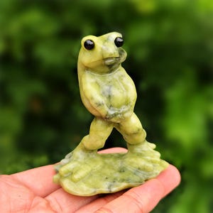 May include: A small, hand-carved frog figurine made from a pale, mottled green stone. The frog is depicted in a standing, upright position with its front limbs resting against its torso, featuring smooth, polished surfaces and two dark, round eyes.