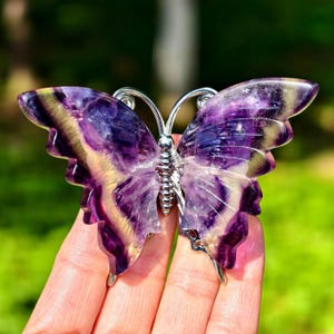 May include: A butterfly-shaped decorative object made of purple and yellow fluorite stone. The butterfly has a silver-colored metal body and antennae. The wings are translucent, showcasing the stone's natural patterns.
