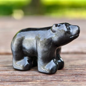 Könnte beinhalten: Kleine, glänzende schwarze Bärenfigur. Der Bär ist mit glatten Linien geschnitzt und hat eine polierte Oberfläche. Die Figur steht aufrecht, wobei alle vier Pfoten sichtbar sind. Der Bär ist ein dekoratives Objekt.