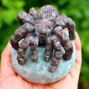 May include: A detailed sculpture of a spider with a textured, brown and gray body, perched on a light blue-green stone. The spider's legs are intricately carved, and the stone has a smooth, rounded shape. The background is a blurred green.