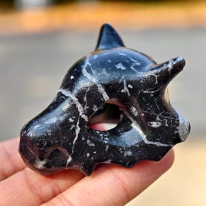 以下が含まれることがあります： 黒と白の大理石の彫刻で、様式化された動物の頭蓋骨です。 頭蓋骨には尖った角、顕著な眉、円形の開口部があります。 彫刻は手で持たれています。