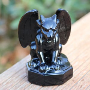May include: A glossy black gargoyle statue with large wings, perched on a hexagonal base. The sculpture has a stern expression and is made of a shiny, reflective material. The statue is a decorative item.