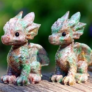 May include: Two small, detailed dragon figurines. The dragons are carved from stone with a mottled pattern of red, green, and brown. They have large eyes and are posed sitting upright. The background is blurred, suggesting an outdoor setting.